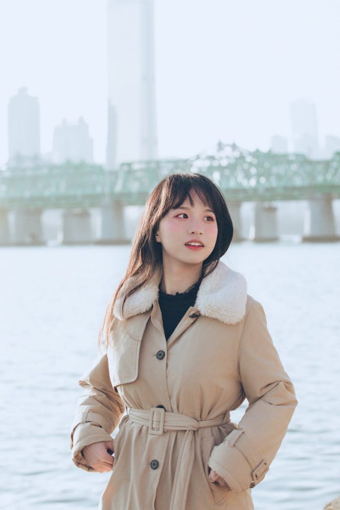 young model in a beige coat with a fur collar standing by the river